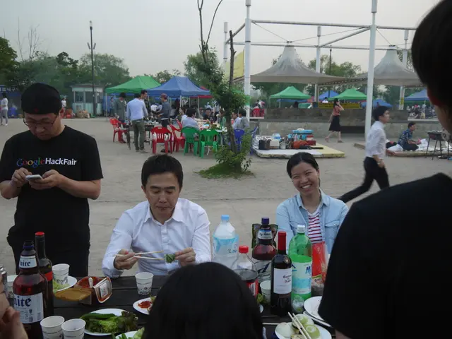 In this image there are group of people. On the table there is a bottle,cup,plate,food. At the back...